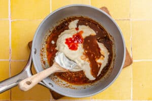 A saucepan with a creamy white sauce, brown sauce, and red sauce being stirred together with a wooden spoon, on a yellow tiled surface.