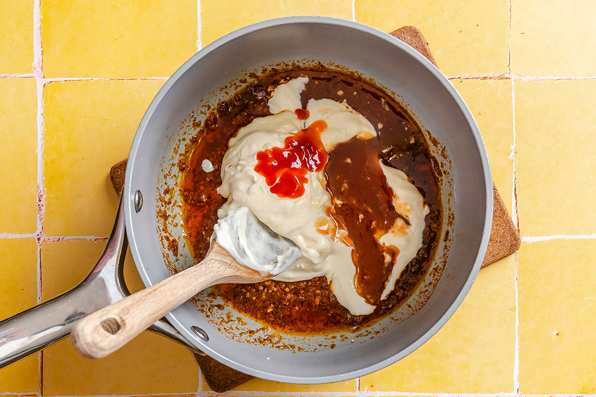 A saucepan with a creamy white sauce, brown sauce, and red sauce being stirred together with a wooden spoon, on a yellow tiled surface.