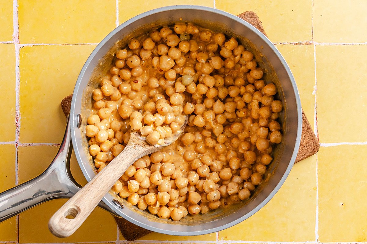 A saucepan filled with cooked chickpeas in a creamy sauce sits on a yellow tiled surface, with a wooden spoon resting inside.