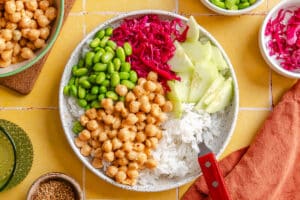 A bowl with white rice, chickpeas, sliced cucumber, edamame, and shredded red cabbage on a yellow tiled surface, with small bowls of toppings nearby.