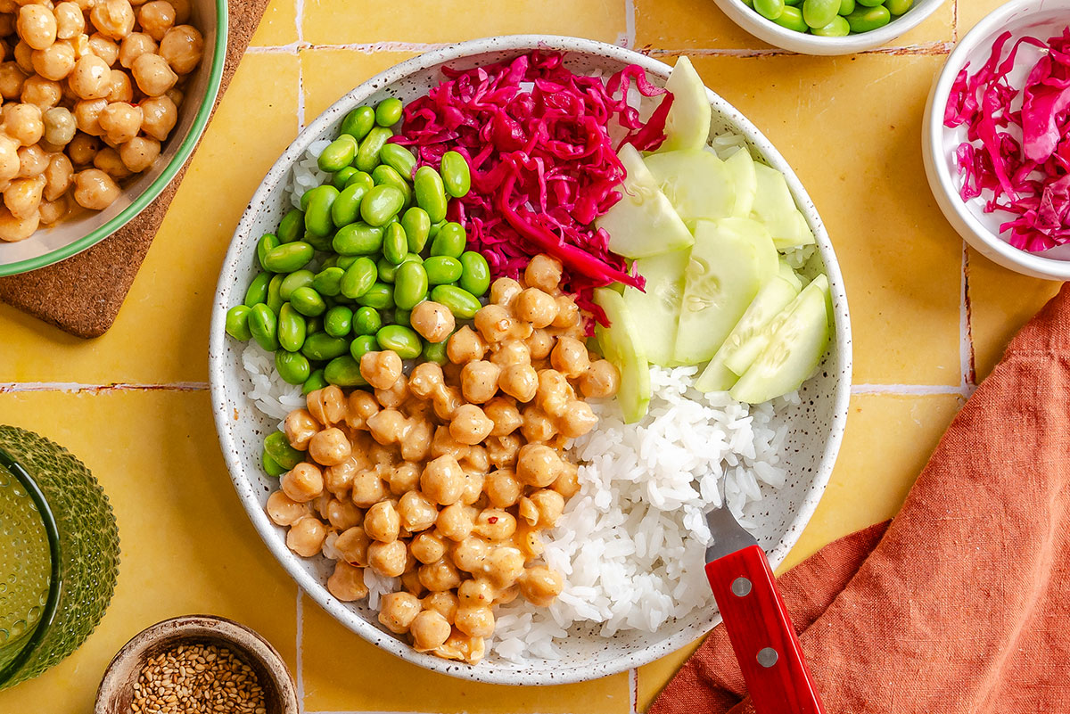 A bowl with white rice, chickpeas, sliced cucumber, edamame, and shredded red cabbage on a yellow tiled surface, with small bowls of toppings nearby.