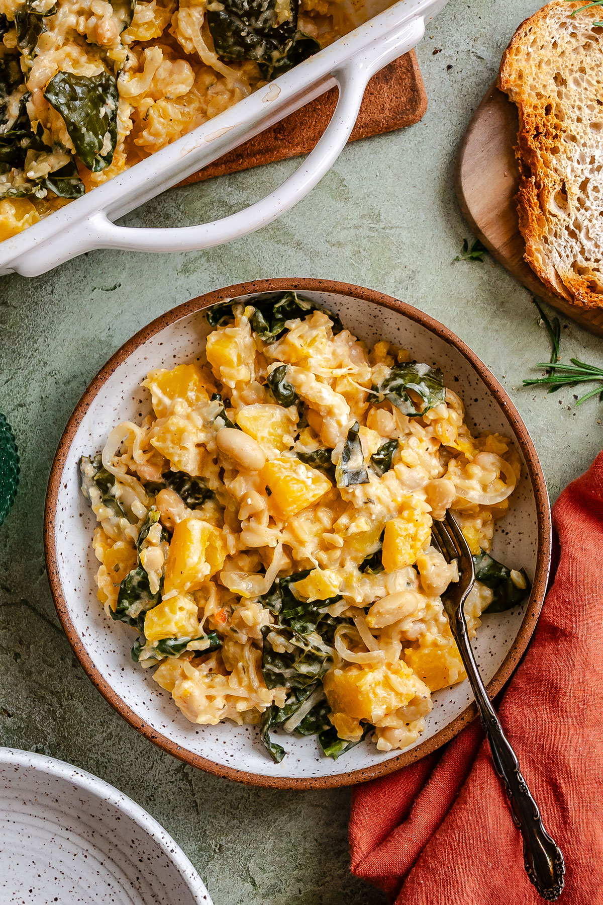 A plate of baked pasta with white beans, kale, and chunks of yellow squash, served with a fork. Sliced bread is beside the plate, and part of a casserole dish is visible.