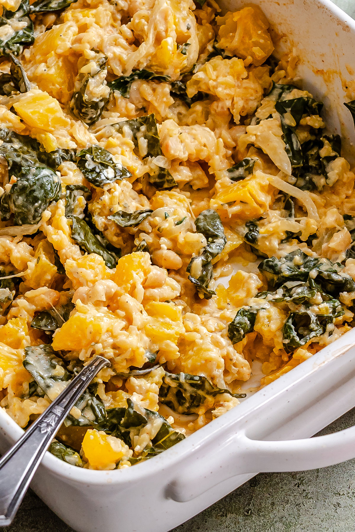 A white baking dish filled with cheesy rice casserole containing chopped greens, yellow bell peppers, and onions, with a serving spoon in the corner.