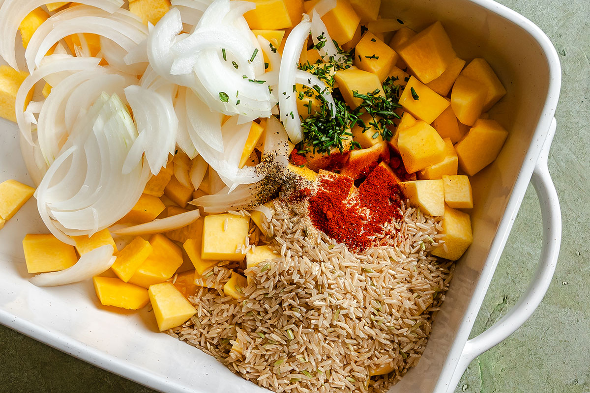 A baking dish containing sliced onions, cubed butternut squash, uncooked brown rice, chopped herbs, black pepper, and paprika.