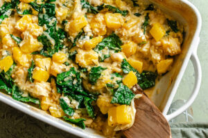 Baked casserole with chunks of yellow squash, leafy greens, white beans, and creamy sauce, mixed together in a white baking dish with a wooden spatula.