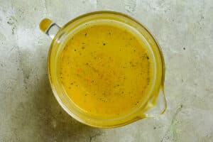 A clear glass measuring cup filled with yellow chicken broth sits on a textured light-colored surface.