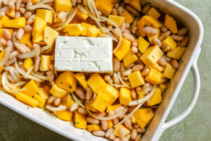 Cubed squash, white beans, sliced onions, herbs, and a block of cream cheese are arranged in a white baking dish, ready to be baked.