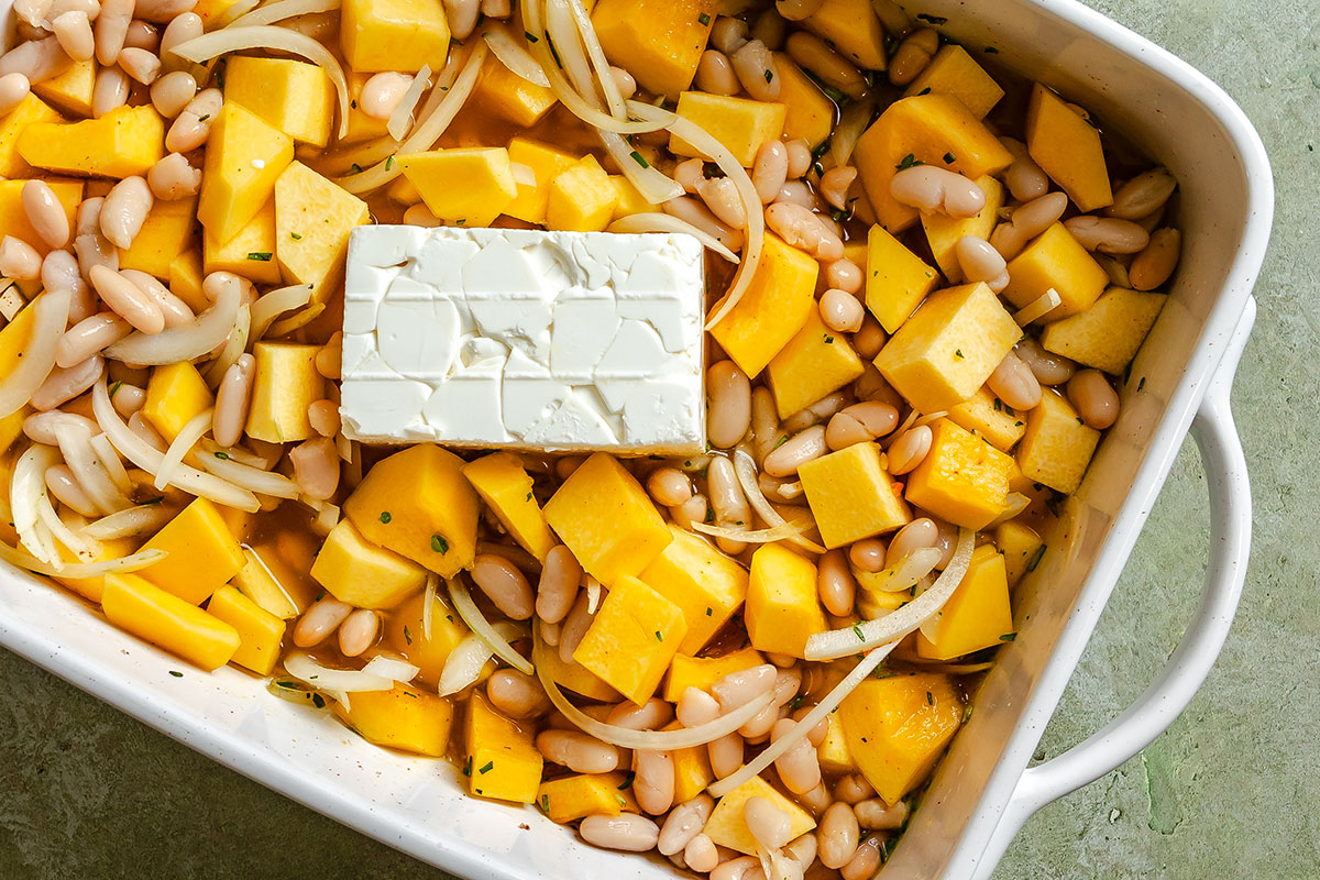 Cubed squash, white beans, sliced onions, herbs, and a block of cream cheese are arranged in a white baking dish, ready to be baked.