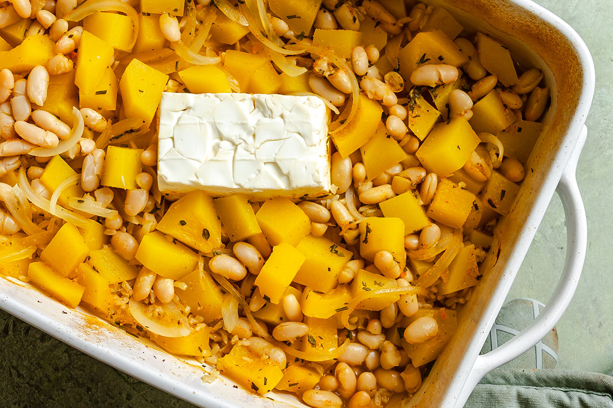 A casserole dish with cubed squash, white beans, sliced onions, herbs, and a block of cream cheese in the center, ready for baking.