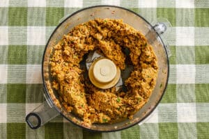 A food processor containing blended chickpea mixture sits on a green and white checkered tablecloth.