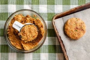 A half-cup measuring cup shapes a veggie patty mixture in a glass bowl; one formed patty is on a parchment-lined baking sheet. Green checkered tablecloth underneath.