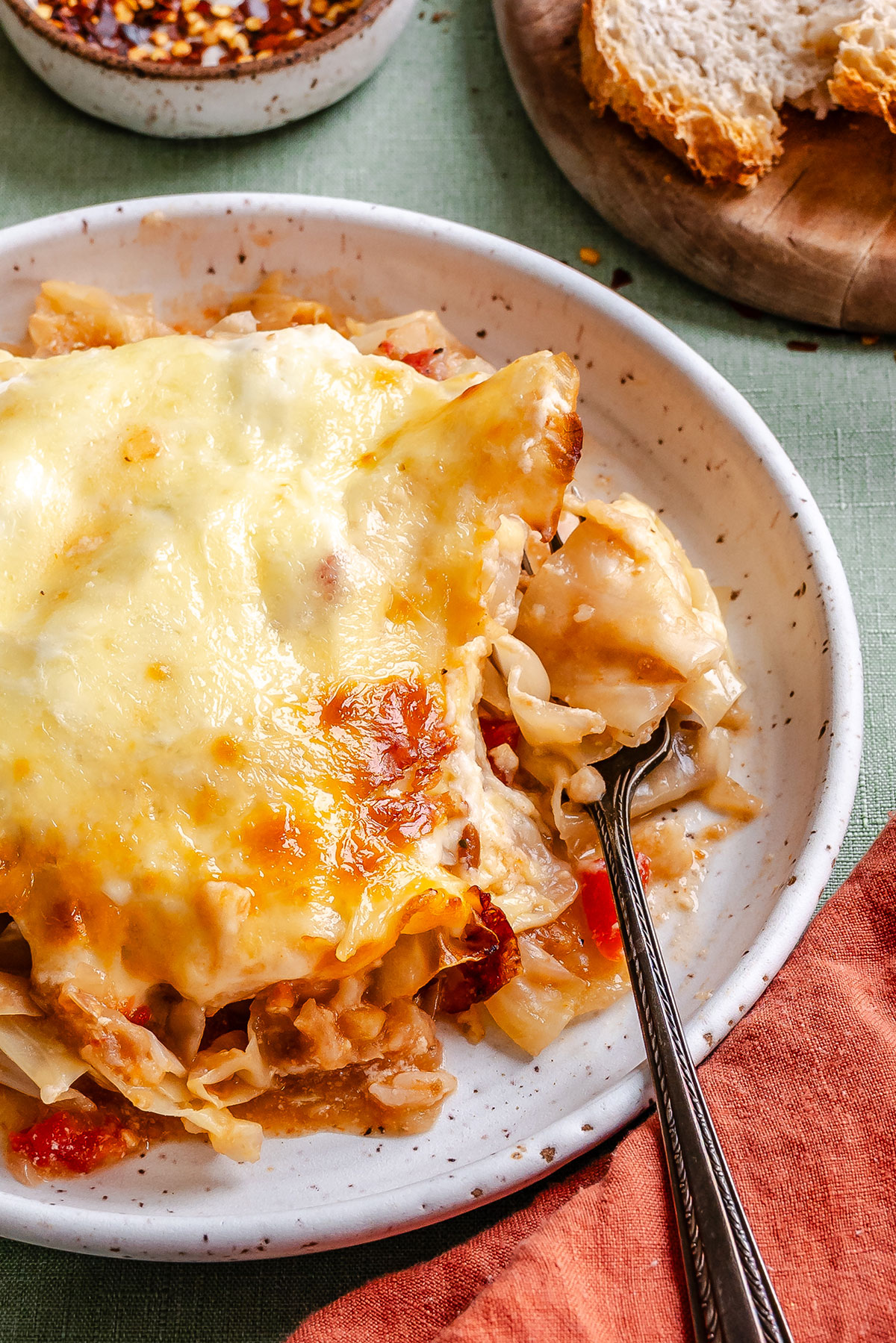 A plate of cheesy baked pasta with a fork, next to a slice of bread and a bowl of red pepper flakes on a green tablecloth.