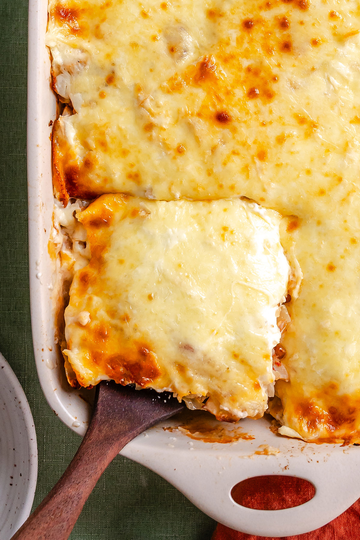 A baked cheesy casserole is being served from a rectangular dish with a wooden spatula.