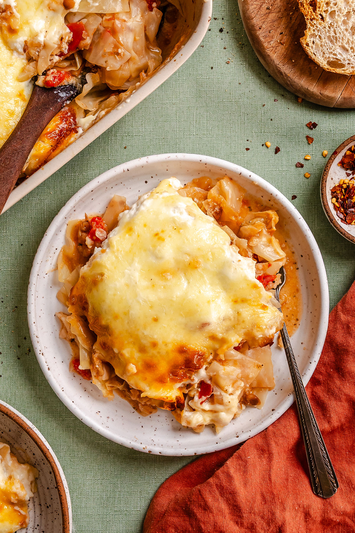 A serving of cheesy baked casserole with visible pasta, sauce, and melted cheese on a white plate, with a fork and bread on the side.