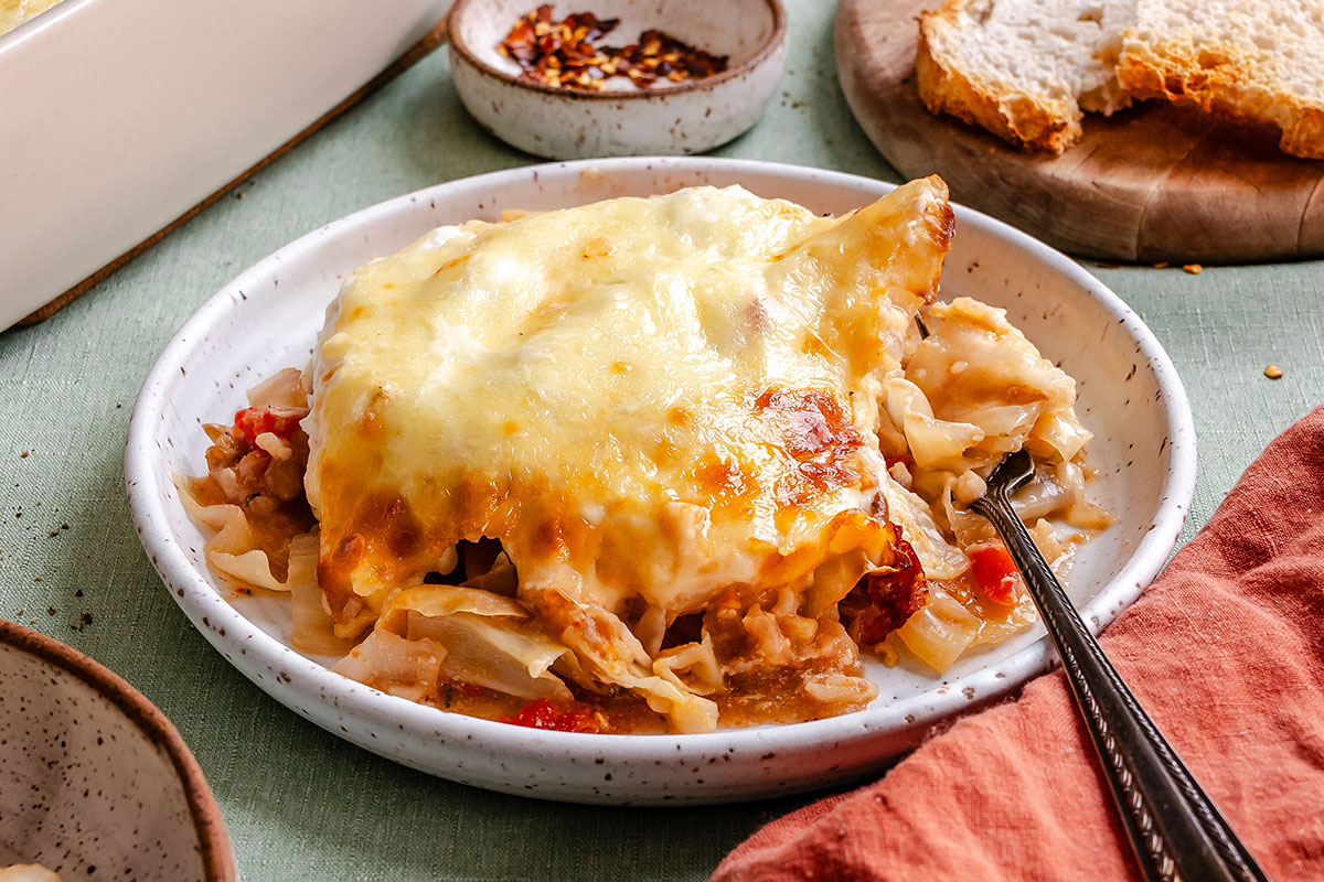 A plate of baked lasagna with melted cheese on top, served with a fork. Bread and a small dish of chili flakes are visible in the background.