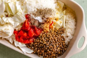A baking dish filled with chopped cabbage, diced tomatoes, lentils, minced garlic, white rice, onions, and various dried herbs and seasonings.