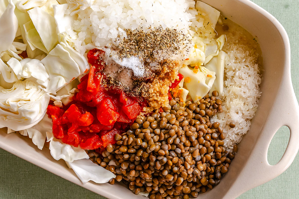 A baking dish filled with chopped cabbage, diced tomatoes, lentils, minced garlic, white rice, onions, and various dried herbs and seasonings.