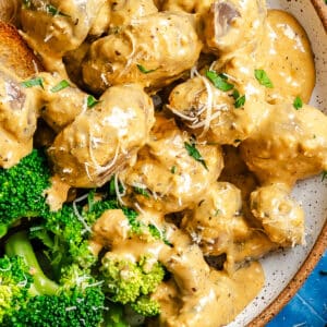 Plate of meatballs in creamy sauce with shredded cheese and herbs, served with steamed broccoli and toasted bread on the side.