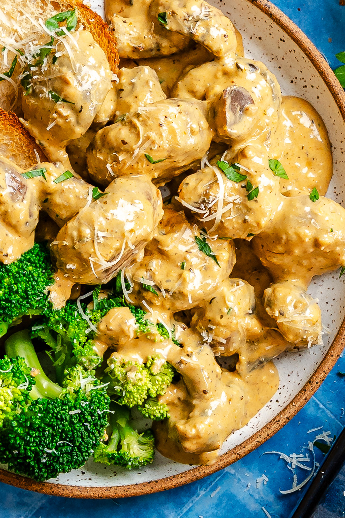 Plate of meatballs in creamy sauce with shredded cheese and herbs, served with steamed broccoli and toasted bread on the side.