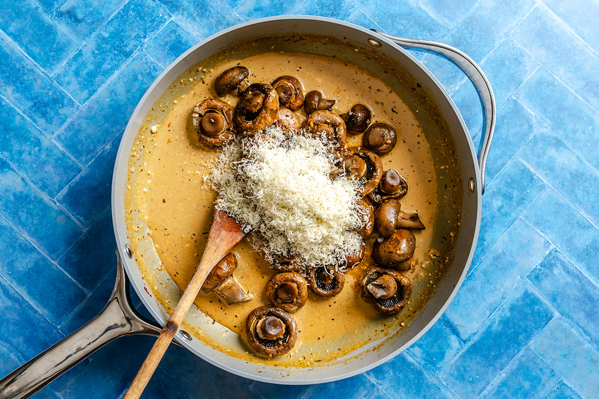 Mushrooms and grated cheese in a creamy sauce are being stirred in a pan with a wooden spoon on a blue tiled surface.