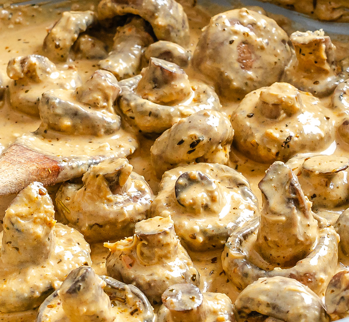 Whole mushrooms coated in a creamy, herb-flecked sauce, with a wooden spoon partially visible, suggesting the dish is being stirred or prepared.