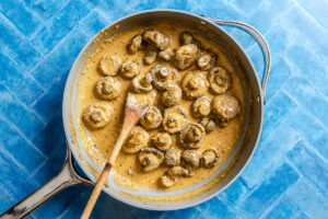 Mushrooms in a creamy sauce are cooked in a skillet with a wooden spoon, placed on a blue tile surface.