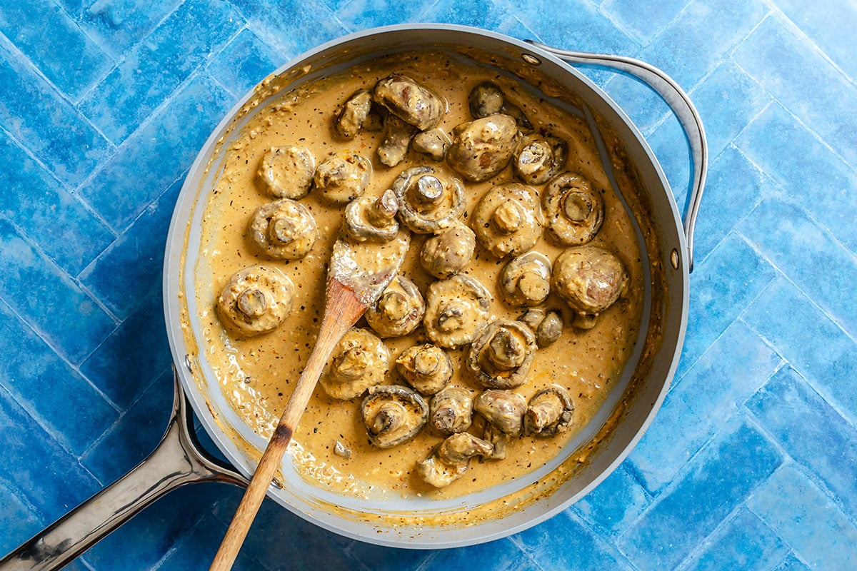 Mushrooms in a creamy sauce are cooked in a skillet with a wooden spoon, placed on a blue tile surface.