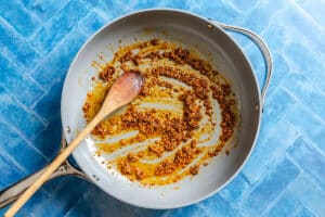 A skillet with browned minced garlic and spices being stirred by a wooden spoon, on a blue tiled surface.