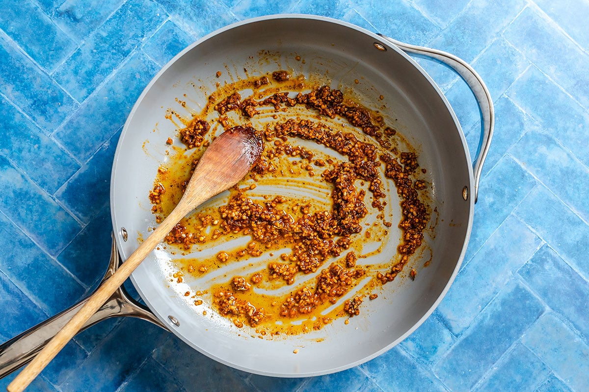 A skillet with browned minced garlic and spices being stirred by a wooden spoon, on a blue tiled surface.