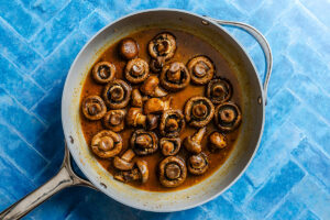 Whole mushrooms sautéed in a brown sauce fill a silver skillet, which rests on a blue tiled surface.