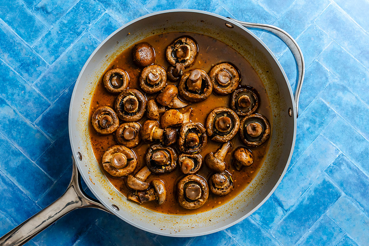 Whole mushrooms sautéed in a brown sauce fill a silver skillet, which rests on a blue tiled surface.