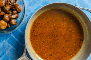 A skillet of brown sauce sits on a blue surface next to a glass bowl filled with cooked mushrooms.