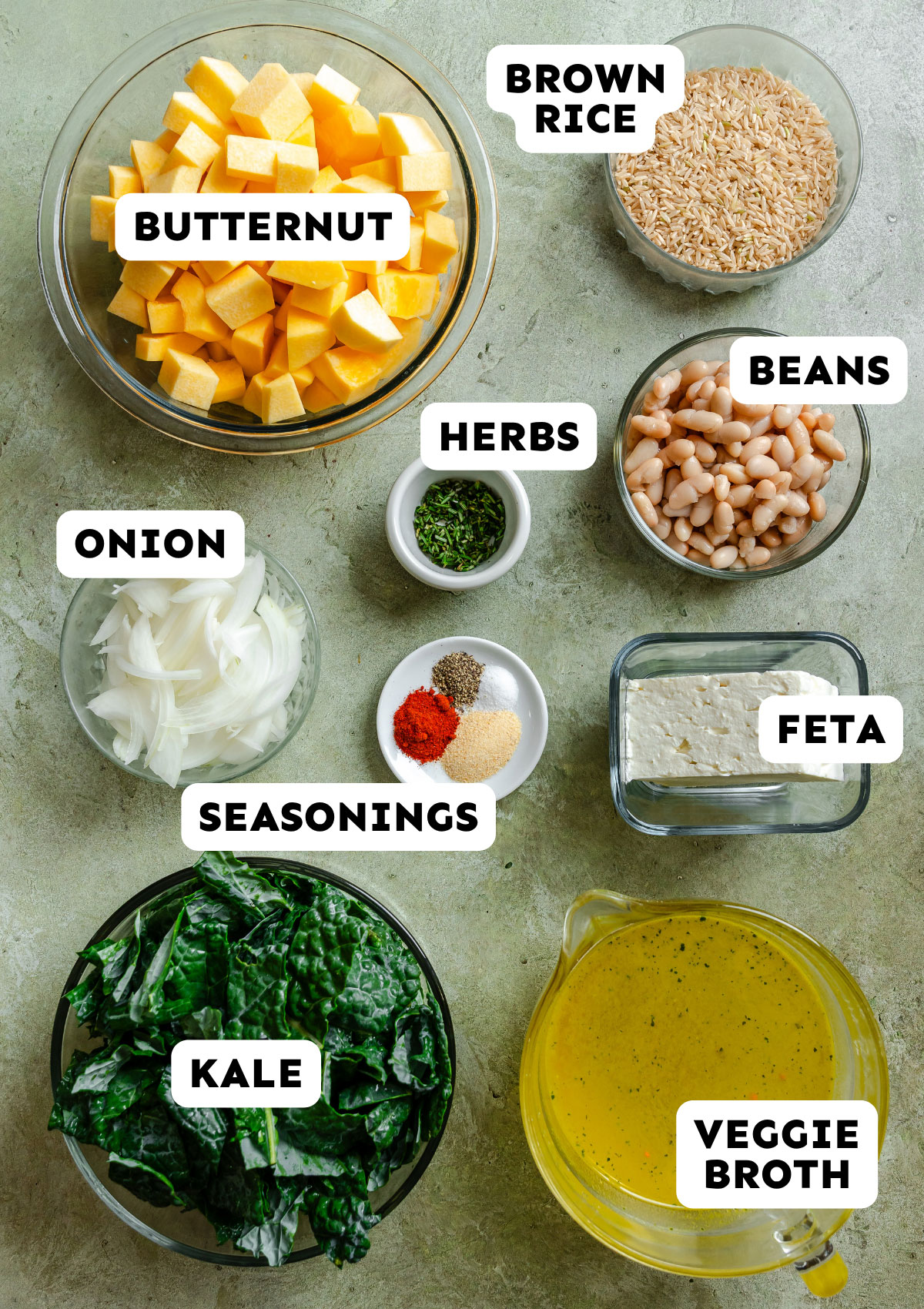 Top-down view of bowls with butternut squash, brown rice, kale, sliced onion, white beans, feta, veggie broth, and seasonings on a light surface—each labeled with text—perfect for assembling an easy butternut squash dump and bake meal.