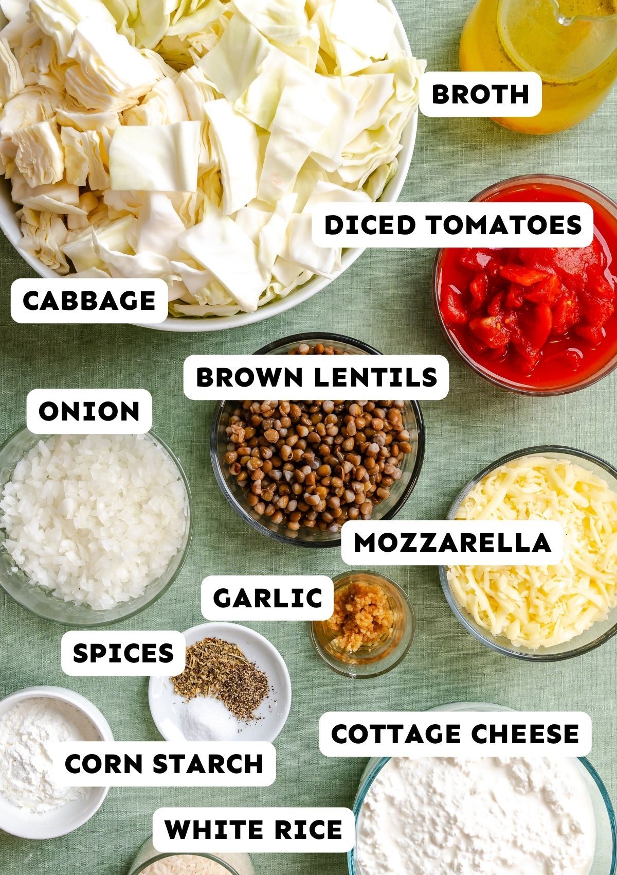 Assorted ingredients on a green surface, labeled: cabbage, onion, garlic, spices, corn starch, white rice, brown lentils, diced tomatoes, broth, mozzarella, and cottage cheese.