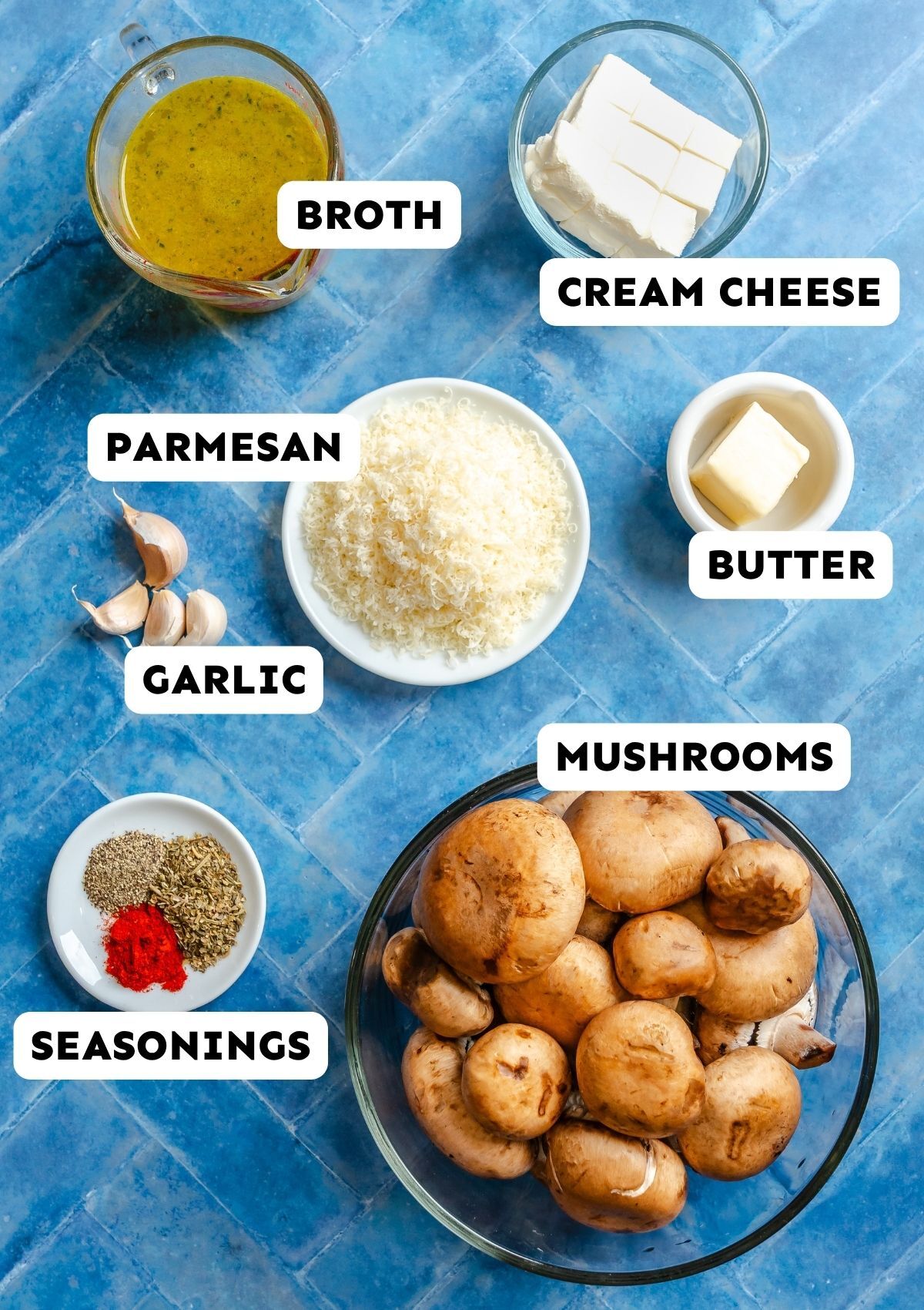 A glass bowl of mushrooms, bowls of grated parmesan, cream cheese, broth, butter, garlic cloves, and a dish of seasonings on a blue tiled surface, each labeled with text.