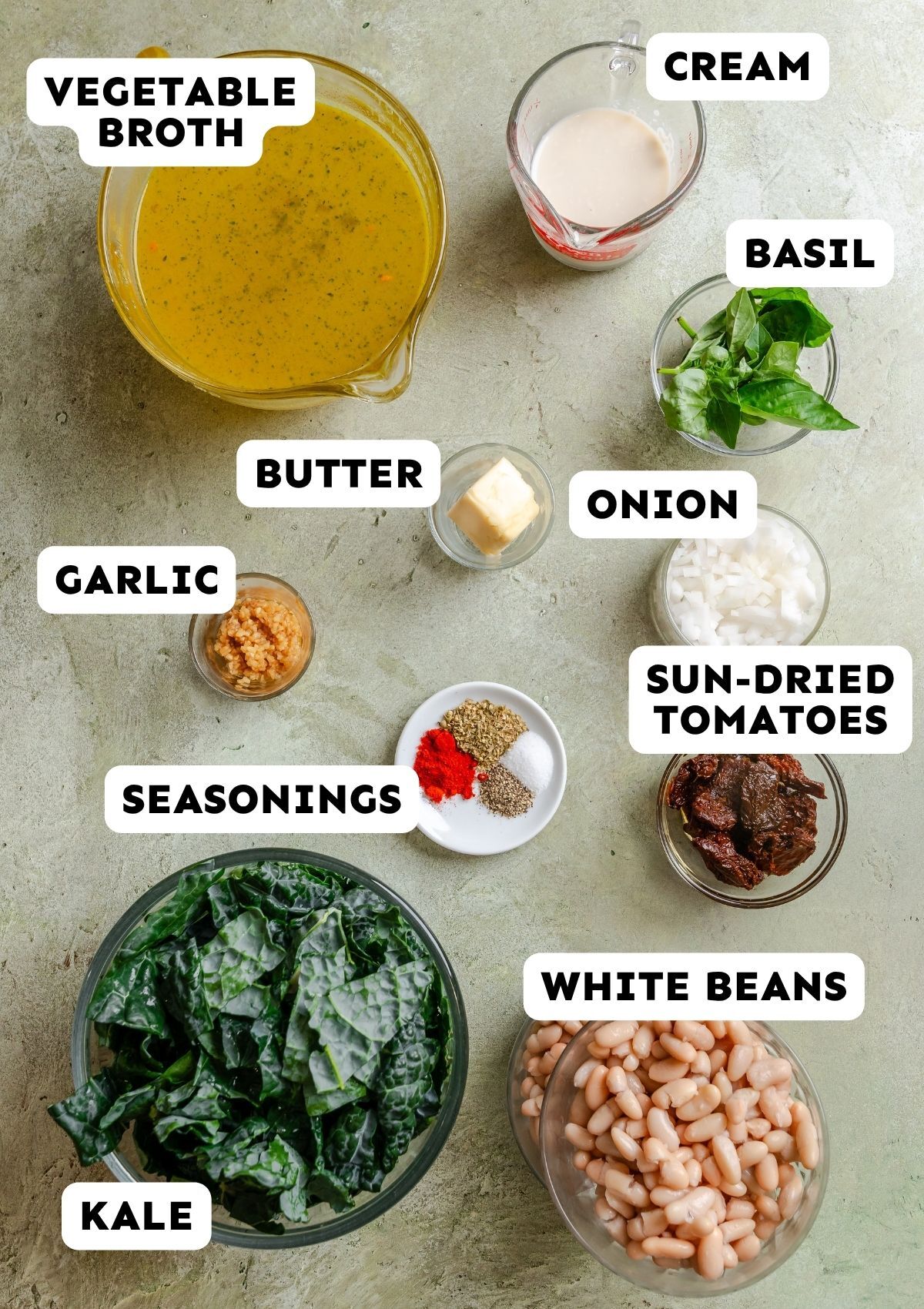 Various soup ingredients in bowls and cups, labeled: vegetable broth, cream, basil, butter, onion, garlic, sun-dried tomatoes, seasonings, kale, and white beans, arranged on a countertop.