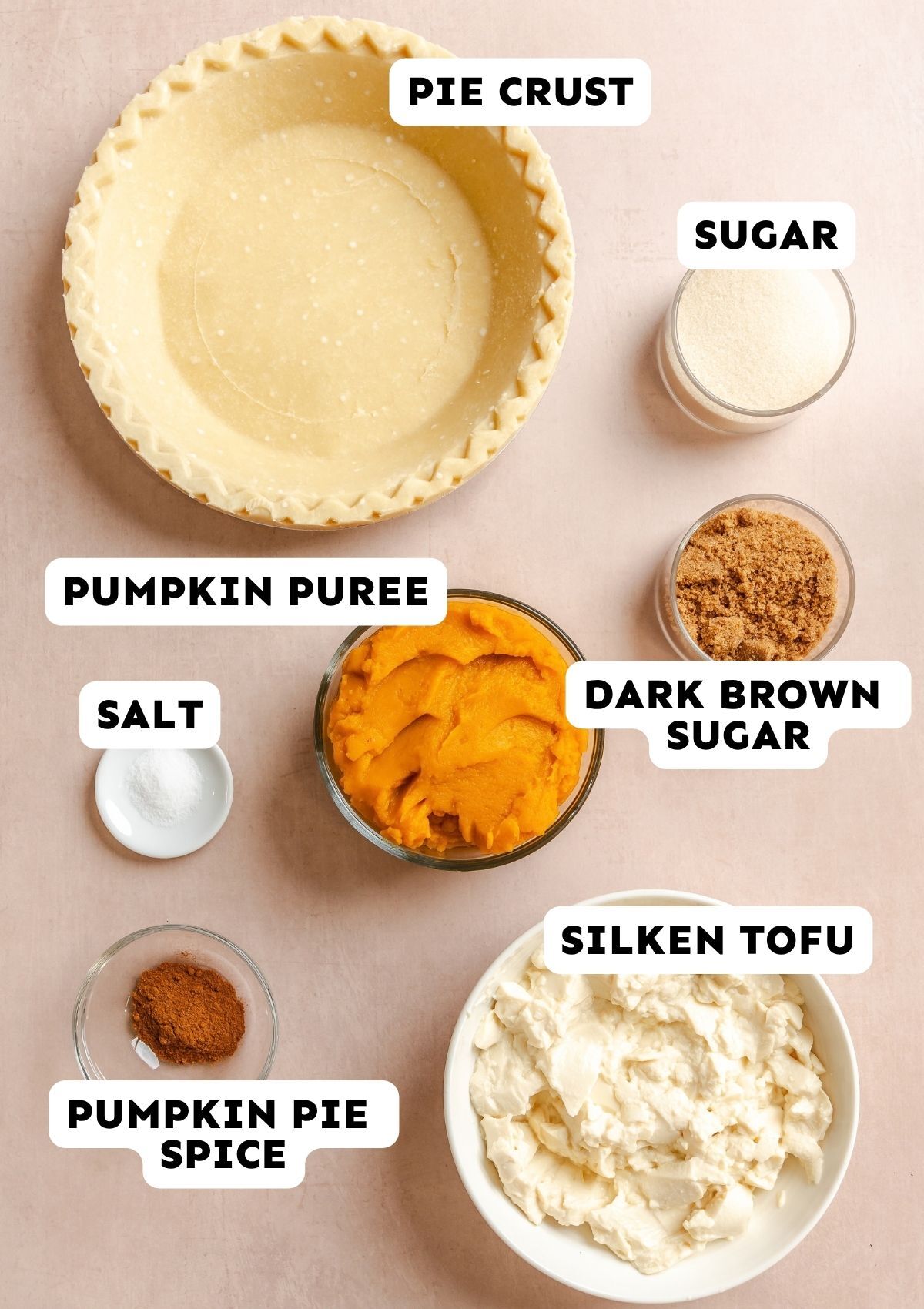 Overhead view of labeled bowls containing pie crust, sugar, dark brown sugar, pumpkin puree, silken tofu, pumpkin pie spice, and salt arranged on a light surface.