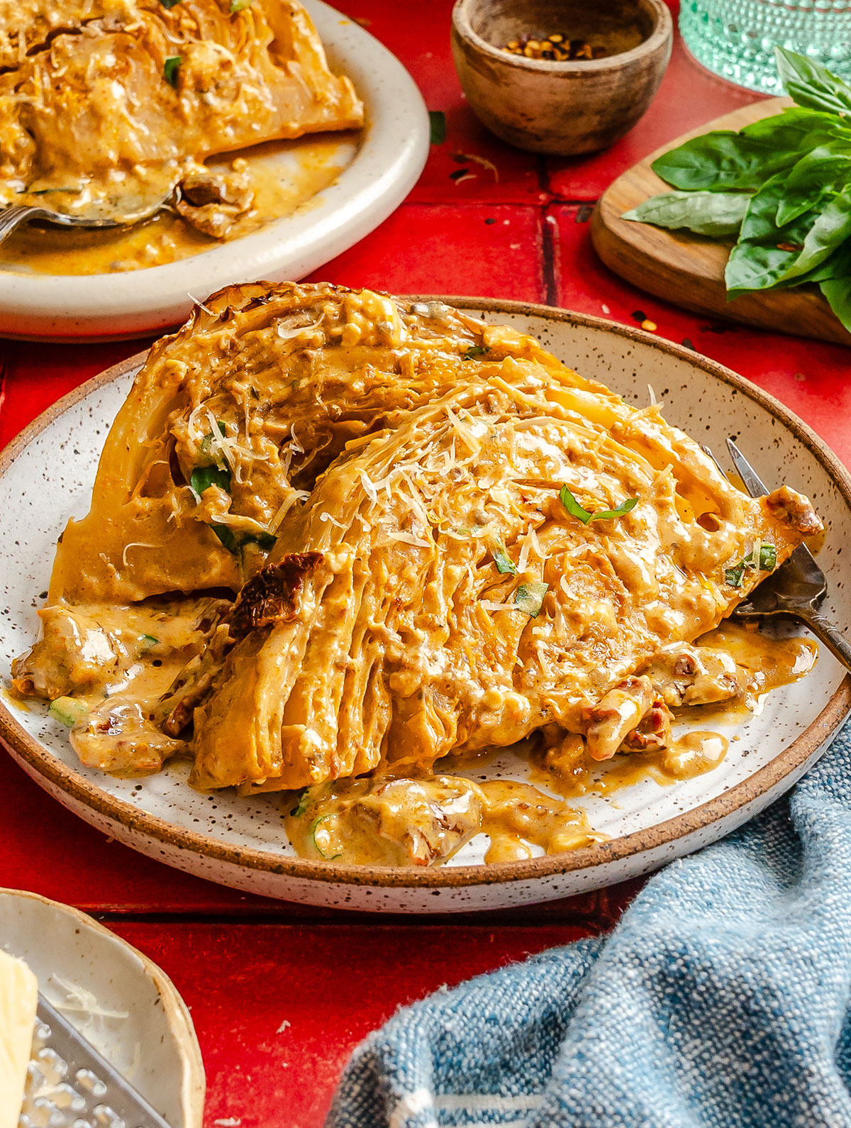 A plate of creamy, golden-brown melting cabbage garnished with fresh herbs, shown on a speckled ceramic dish with a fork, against a red tabletop background.