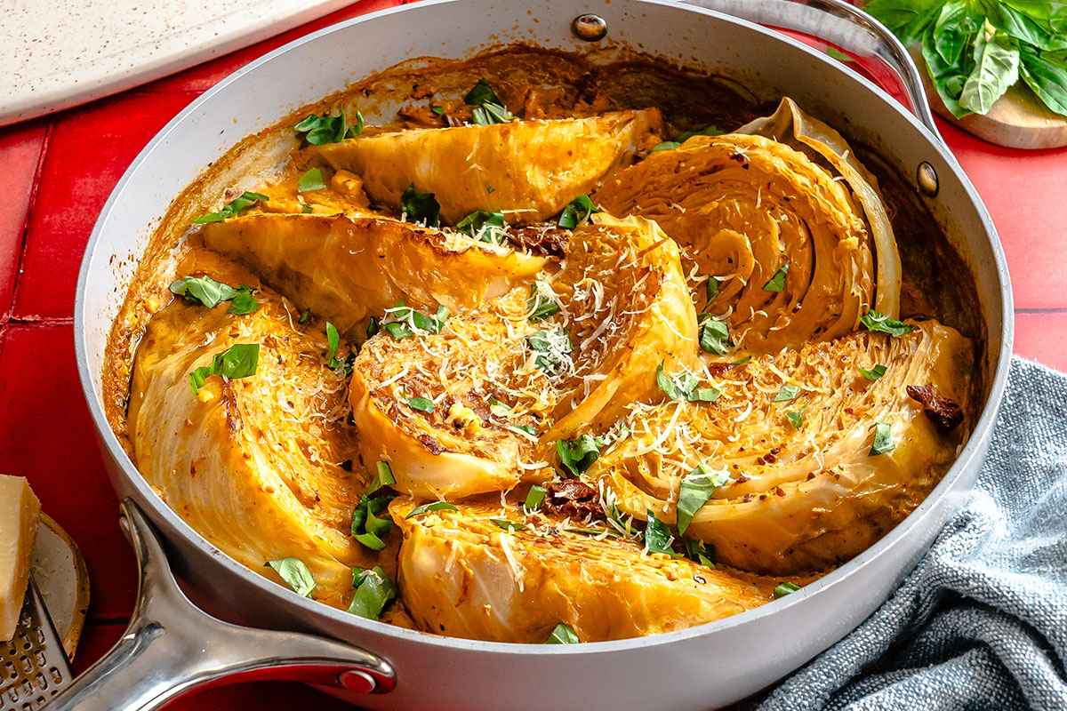 Wedges of cabbage in a creamy sauce are cooked in a skillet and garnished with grated cheese and chopped herbs.