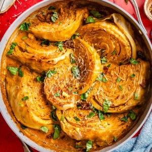 Sautéed cabbage wedges in a creamy sauce, garnished with grated cheese and fresh herbs, served in a skillet on a red tiled surface.