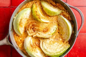 Large cabbage wedges simmer in a skillet filled with creamy, seasoned sauce, set on a red tiled surface.