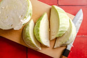 A cabbage cut into quarters sits on a wooden cutting board next to a knife, with a red tiled surface underneath.