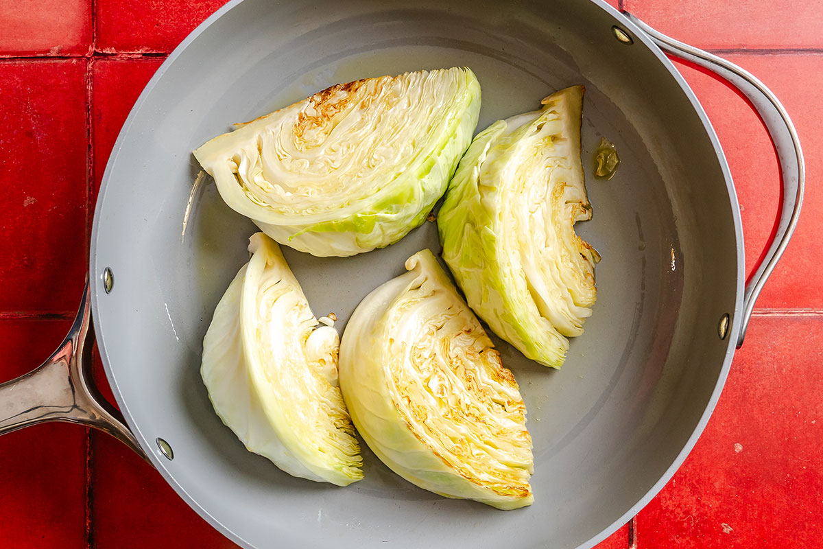 Four wedges of cabbage are being seared in a large gray skillet on a red tiled surface.