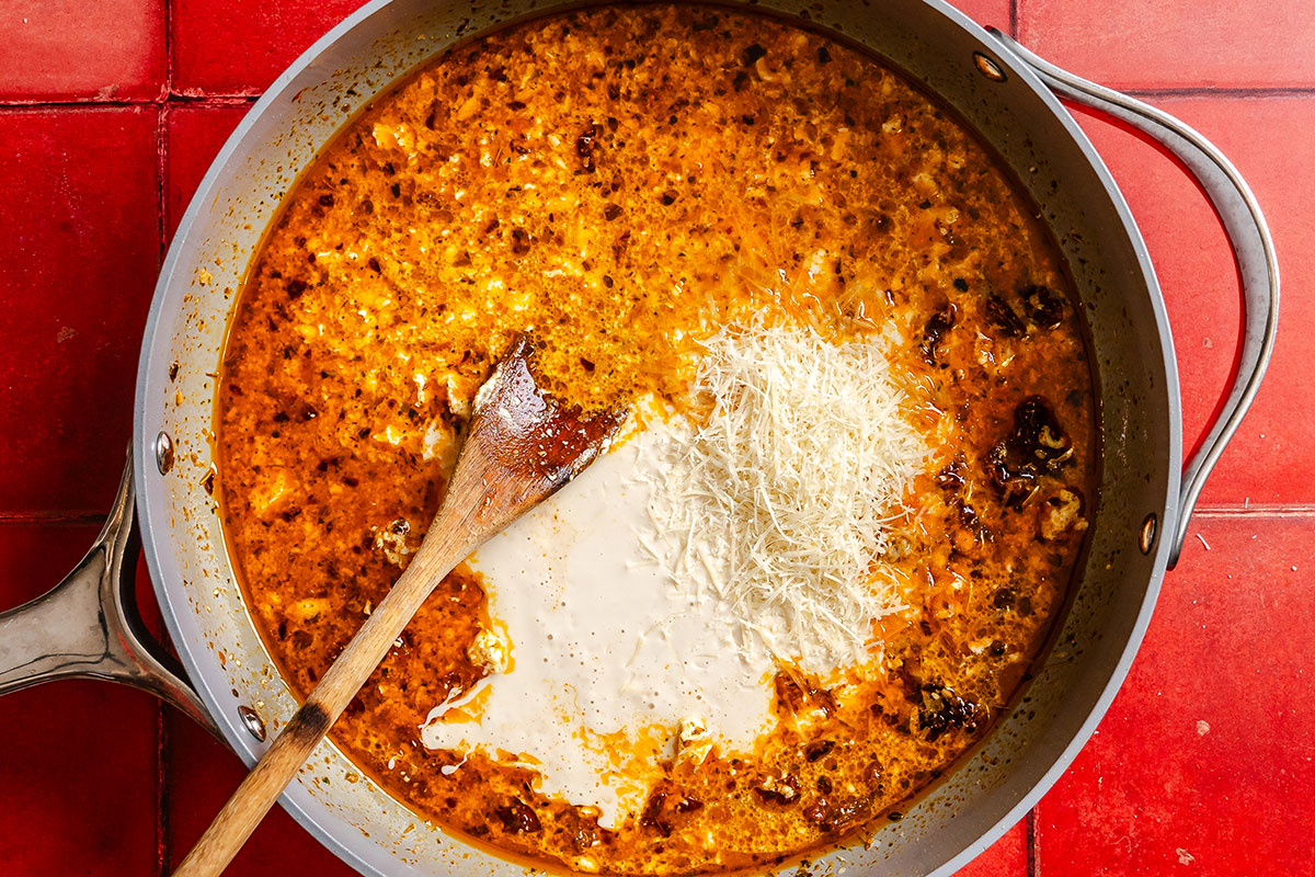 Large skillet with red sauce, cream, shredded cheese, and a wooden spoon on a red tiled surface.