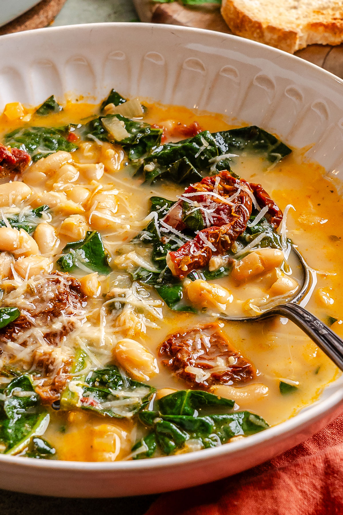 A bowl of white bean and kale soup with sun-dried tomatoes and grated cheese, with a spoon resting inside the bowl.