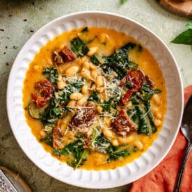 A white bowl filled with bean and kale soup, topped with sun-dried tomatoes and grated cheese, sits on a green surface next to bread and a spoon.