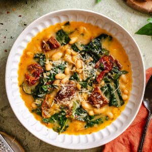 A white bowl filled with bean and kale soup, topped with sun-dried tomatoes and grated cheese, sits on a green surface next to bread and a spoon.