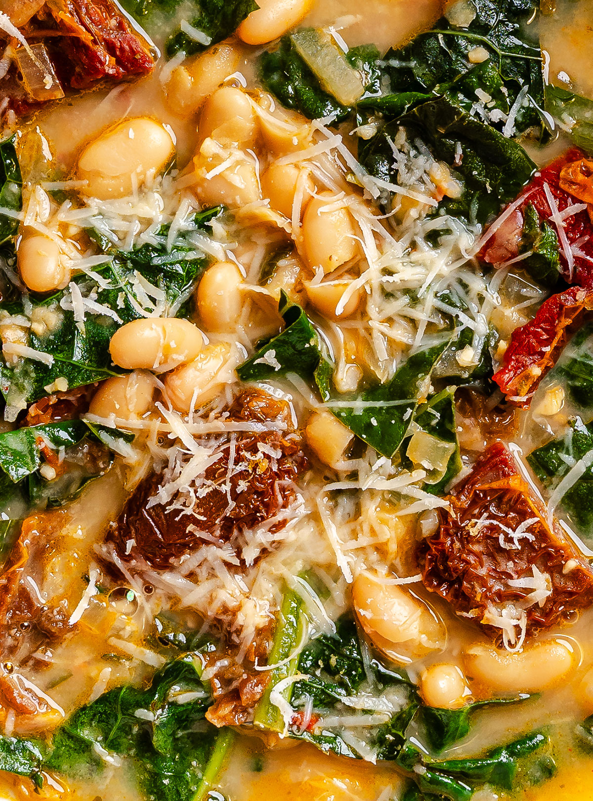 Close-up of a soup with white beans, shredded greens, sun-dried tomatoes, and grated cheese on top.