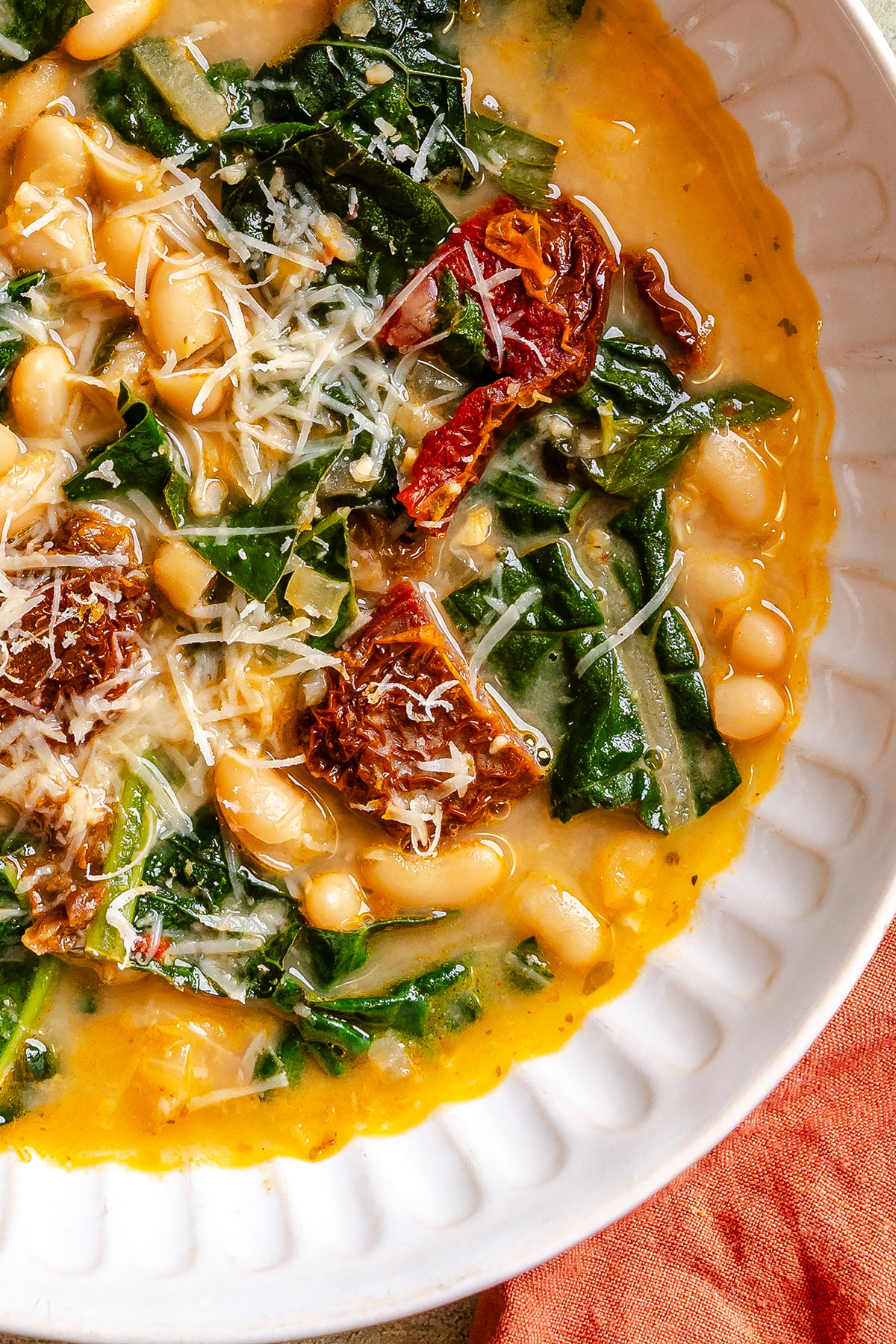 A bowl of creamy soup with white beans, chopped greens, sun-dried tomatoes, and grated cheese in a white dish.