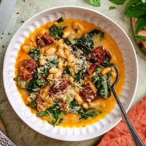 A bowl of white bean and kale soup topped with grated cheese and sun-dried tomatoes, with a spoon in the bowl and bread, basil, and cheese nearby.